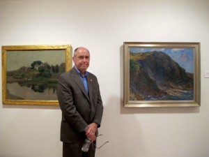 Ogunquit Museum of Art director Ron Crusan stands between The Cliff by Charles Woodbury, and First Bridge, Perkins Cove by Hamilton Easter Field.