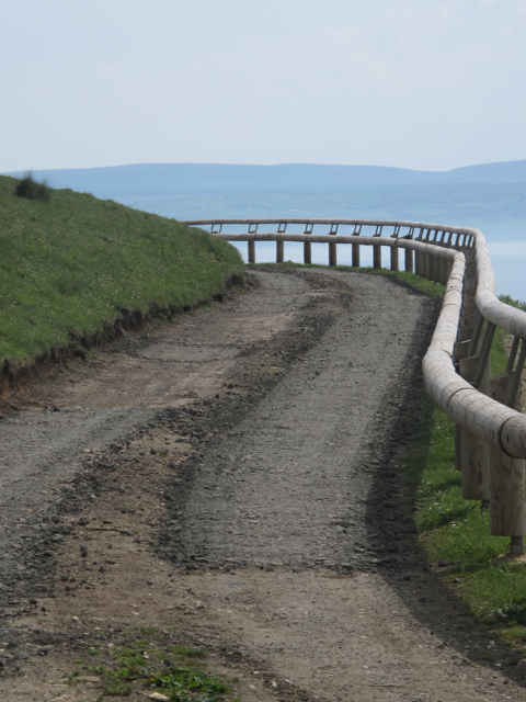 A fence and a hillside frame a narrow lane with a sharp bend is a reminder to keep left when driving in Ireland